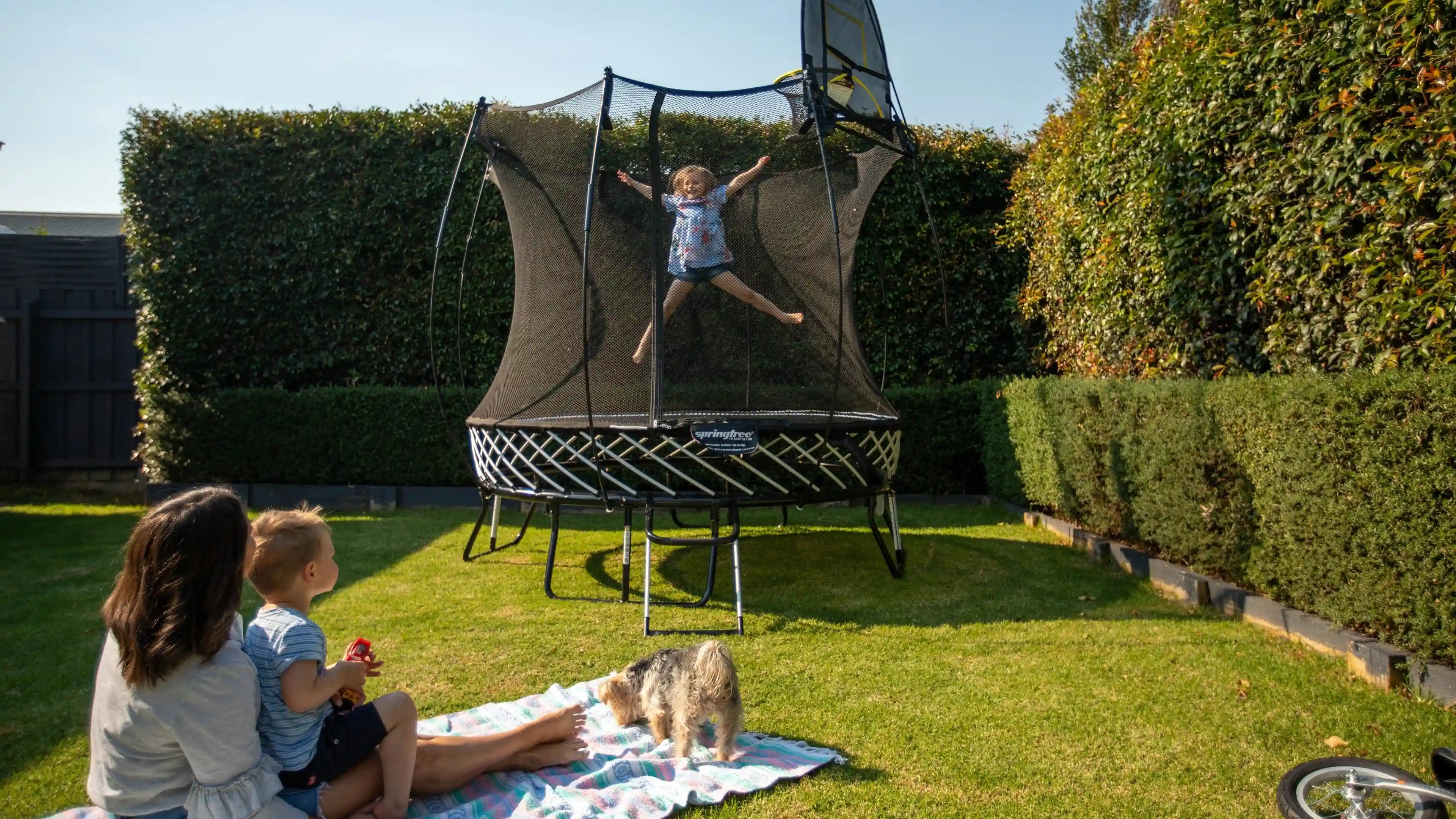 girl jumping so high on an outdoor trampoline while a lady and a kid are watching her