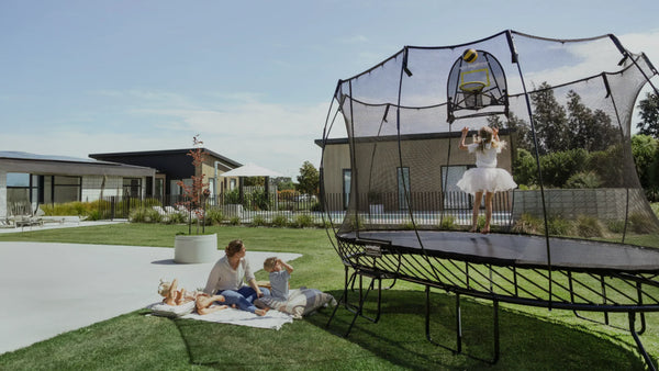 A little girl jumping on a Springfree Trampoline while a young boy and woman sit outside on a blanket next to the trampoline.
