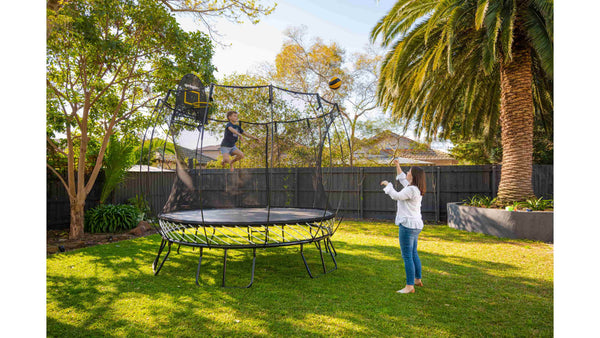 A mom throwing a ball to her son who's jumping on a trampoline.