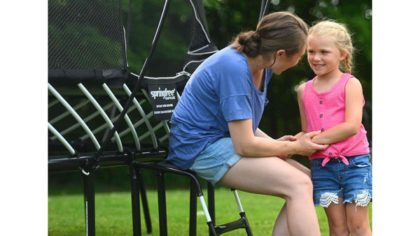 A giddy mother sitting on a trampoline ladder holding her smiling daughter's hands. 