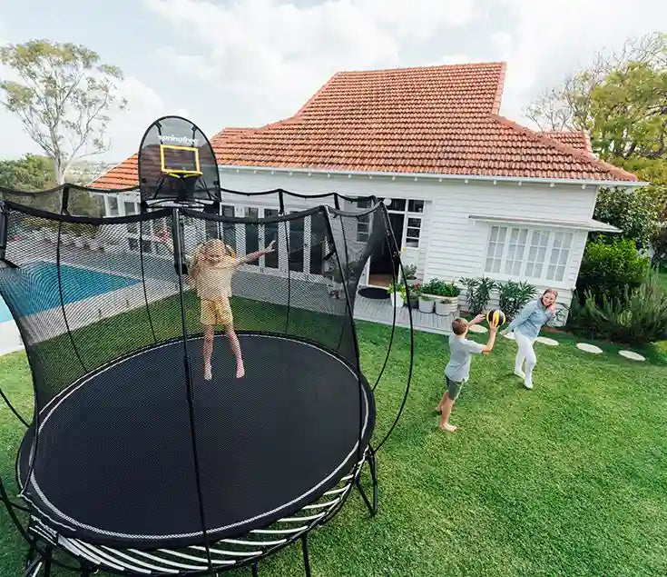 girl jumping on a trampoline while a boy and a lady is playing basketball outside