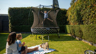 Load image into Gallery viewer, girl jumping so high on an outdoor trampoline while a lady and a kid are watching her

